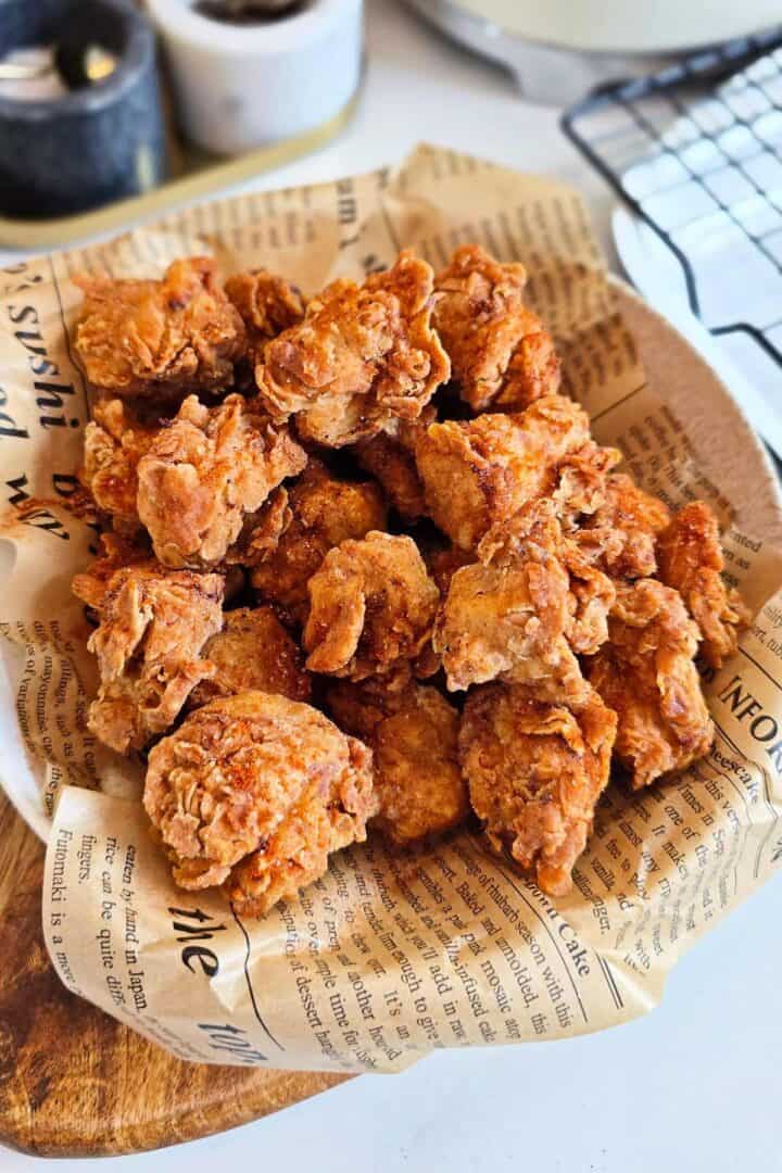 Spicy popcorn chicken sprinkled with spice dust, piled on newspaper-lined paper on a wooden board.