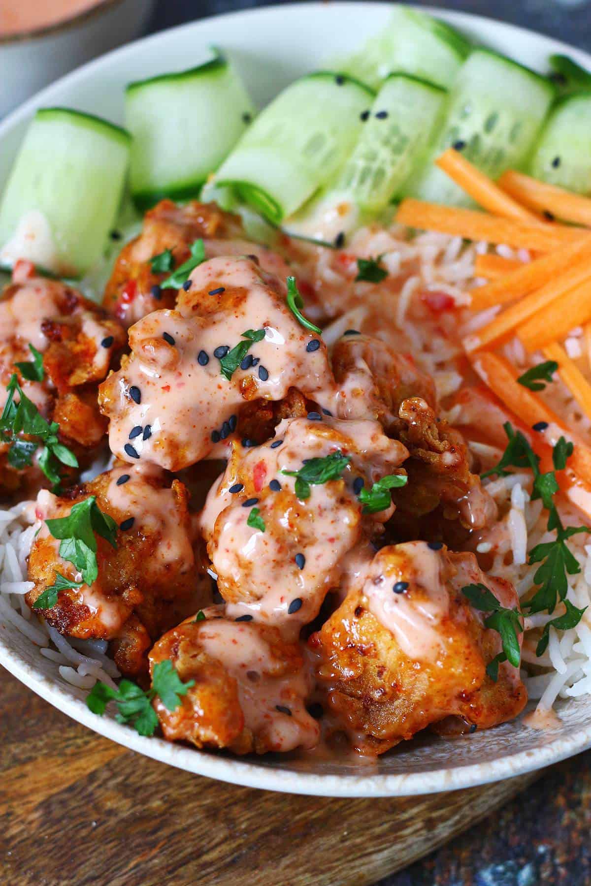 Close-up of crispy fried chicken pieces coated in creamy bang bang sauce, topped with black sesame seeds and parsley, with cucumber ribbons and carrot sticks behind.