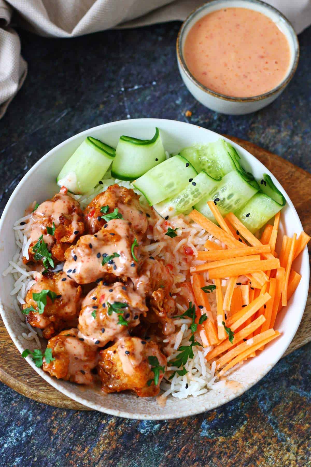 Overhead view of a bang bang chicken bowl with rice, cucumber ribbons and carrot sticks, with a small bowl of sauce on the side.