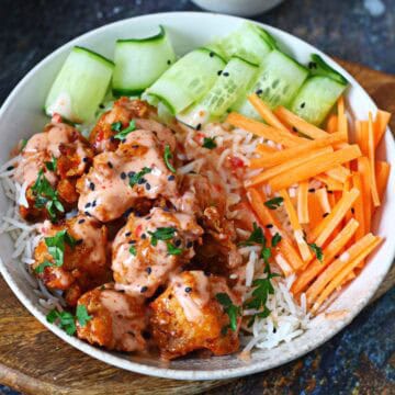 Bang bang chicken bowl with saucy fried chicken over steamed rice, cucumber ribbons and carrot sticks on a wooden board.