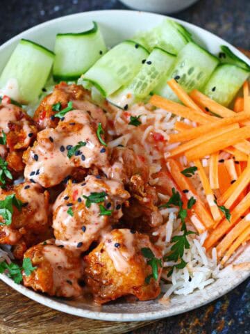 Bang bang chicken bowl with saucy fried chicken over steamed rice, cucumber ribbons and carrot sticks on a wooden board.