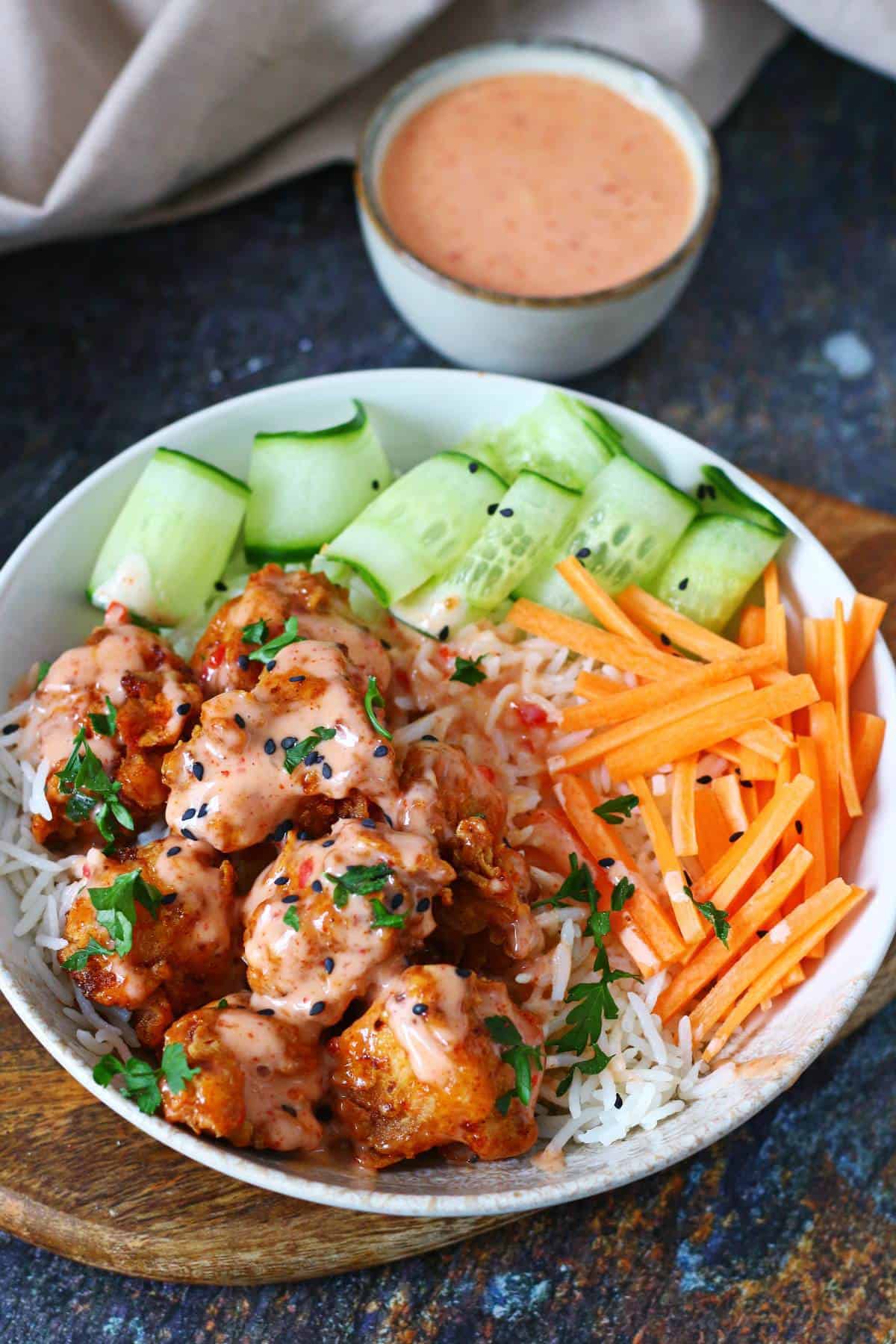 Fried chicken drizzled with sweet chili sriracha sauce in a rice bowl with cucumber and carrots, and extra sauce in a small bowl behind.