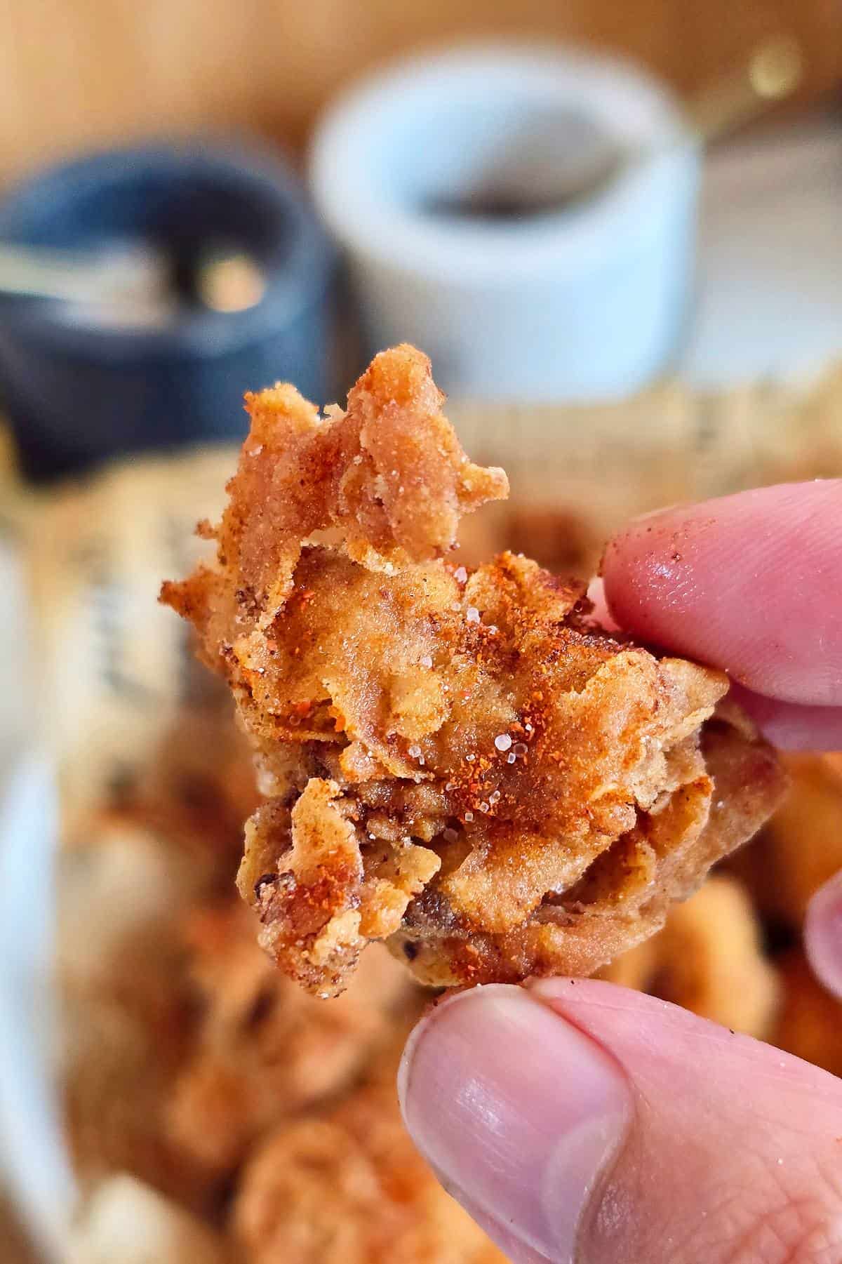 Close-up of a piece of spicy popcorn chicken held between fingers, showing the flaky, crispy coating and spice dust.