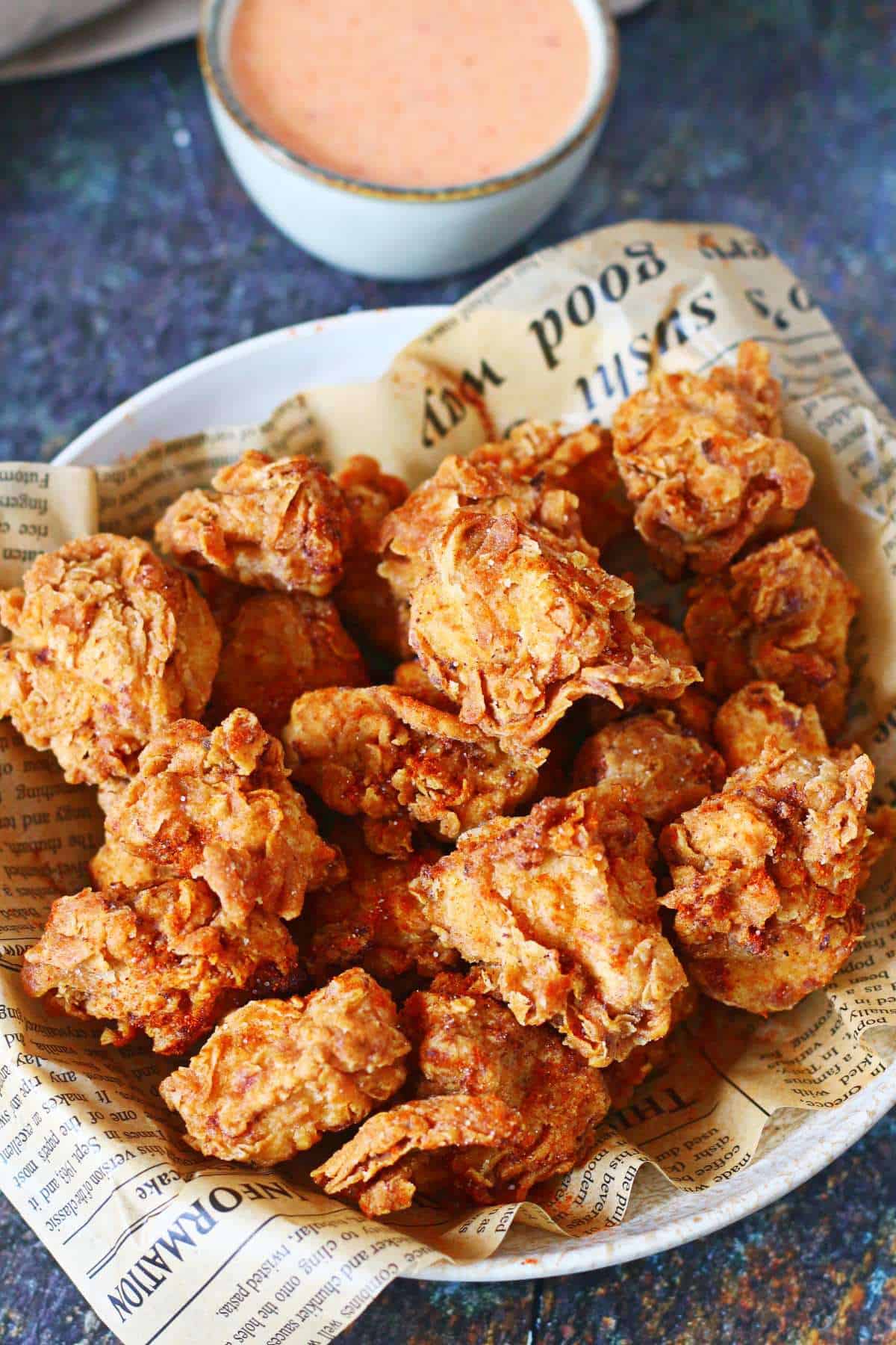 Spicy popcorn chicken in a newspaper-lined bowl with a creamy dipping sauce on a dark background.