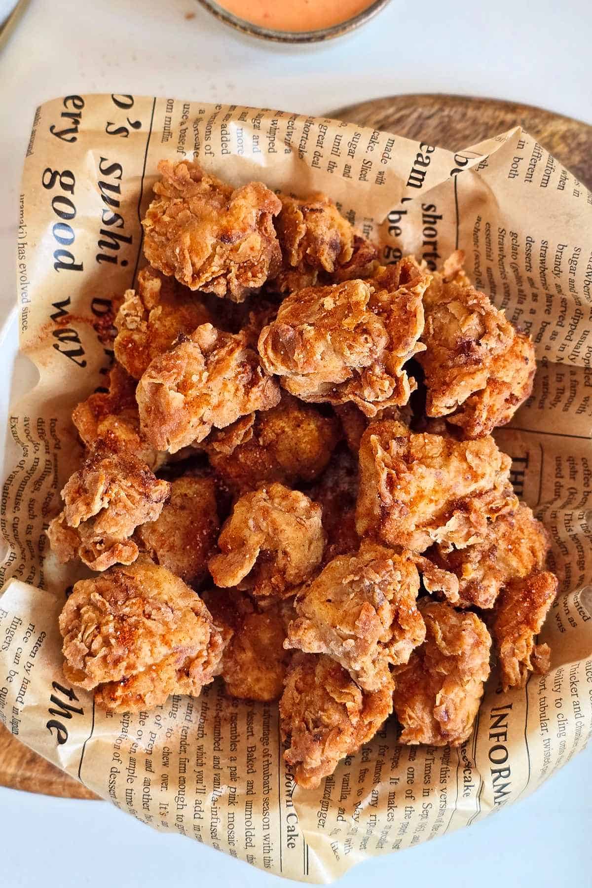 Overhead view of crispy spicy popcorn chicken in a newspaper-lined bowl on a wooden board.
